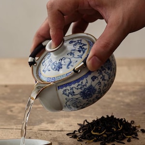 Op de afbeelding: Een wit en blauw porseleinen theepot met een bloemmotief giet water in een kopje. De theepot heeft een zwart handvat en gouden rand. Een hoop losse theeblaadjes ligt in de buurt.