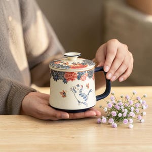 May include: Cream-colored ceramic mug with a lid, featuring a blue and white cat design and floral patterns. The mug has a dark blue handle and a matching lid with a small knob. Butterflies are also depicted on the mug.