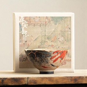 May include: A ceramic bowl with a unique design, featuring a black and gold kintsugi repair and red leaf accents. The bowl is set in front of a box with a traditional Asian-style painting. The bowl has a black base.