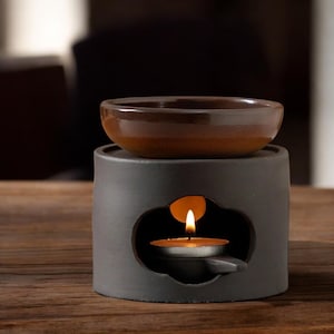 Puede incluir: Un difusor de aceites esenciales de cerámica gris oscuro con un cuenco marrón en la parte superior. Una vela de té encendida está dentro del difusor, visible a través de una abertura decorativa. El difusor está sobre una superficie de madera.