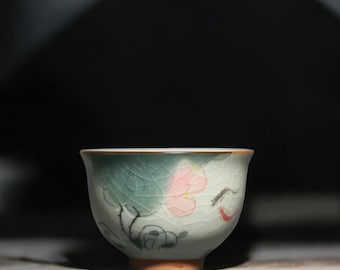 May include: A ceramic tea bowl with a crackle glaze in shades of green and white. The bowl features a pink floral design with black and red accents. The rim has a brown trim. The bowl is set on a wooden surface.
