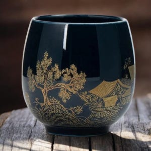Puede incluir: Una taza de té de cerámica azul oscuro con un diseño dorado de paisaje que presenta un árbol y edificios. La taza tiene una forma redondeada y un acabado brillante, adecuada para té u otras bebidas.