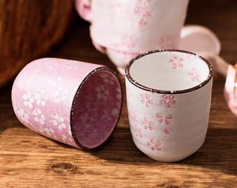 May include: Two pink ceramic cups with a floral pattern. One cup is lying on its side, while the other is upright. The cups have a dark brown rim and are set on a wooden surface. A third cup is visible in the background.