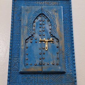 Könnte beinhalten: Ein rechteckiger blauer Wandbehang aus Holz in Form einer Doppeltür mit Rundbogen. Der Rahmen und die Türen weisen aufwendige geschnitzte geometrische Muster auf. Die Türen sind mit silberfarbenen Metallnieten und einem goldenen Riegel verziert.