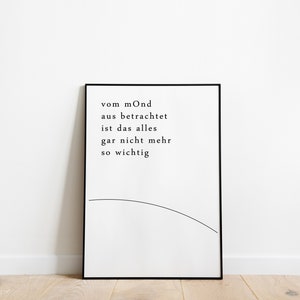 Könnte beinhalten: Schwarz gerahmter Druck mit deutschem Text: „vom mOnd aus betrachtet ist das alles gar nicht mehr so wichtig“. Das minimalistische Design beinhaltet eine geschwungene Linie. Der Druck steht vor einer weißen Wand auf einem Holzboden.