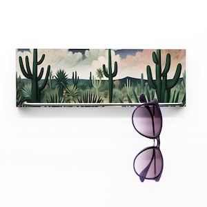 Könnte beinhalten: Ein rechteckiger, an der Wand befestigter Sonnenbrillenhalter mit einem Wüstenlandschaftsdesign mit Kakteen und anderen Wüstenpflanzen. Eine lila Sonnenbrille hängt an der Metallstange. Der Hintergrund ist weiß.