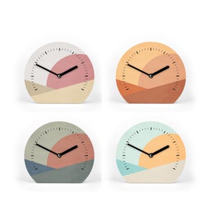 Könnte beinhalten: Vier dekorative analoge Uhren mit abstrakten Landschaftsdesigns. Jedes Zifferblatt hat eine andere Farbpalette, darunter Rosa-, Orange- und Blautöne. Die Uhren haben schwarze Zeiger und ein minimalistisches Design, perfekt für ein modernes Zuhause.
