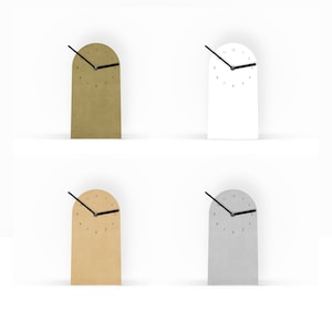 Könnte beinhalten: Vier minimalistische Schreibtischuhren in Bogenform in Gold, Weiß, Beige und Silber. Jede Uhr hat einen schwarzen Zeiger und ein einfaches, nummeriertes Zifferblatt. Die Uhren werden vor einem weißen Hintergrund präsentiert, der ihr modernes Design hervorhebt.