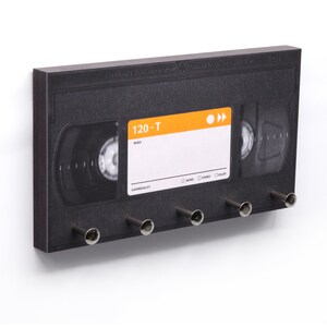 Könnte beinhalten: Ein schwarzer Schlüsselhalter in Form einer VHS-Kassette mit fünf silbernen Haken. Das Etikett trägt die Aufschrift "120-T" und "SUPERQUALITY". Das Design erinnert an klassische Videokassetten.