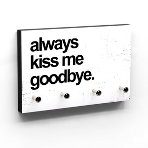 Könnte beinhalten: Ein rechteckiger, weißer Schlüsselhalter mit dem Text "always kiss me goodbye" in Schwarz. Der Schlüsselhalter hat einen schwarzen Rand und vier silberne und schwarze Knöpfe zum Aufhängen von Schlüsseln.