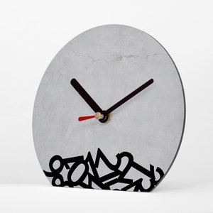 Könnte beinhalten: Eine moderne, graue Uhr mit schwarzen Zeigern und einem roten Sekundenzeiger. Das Zifferblatt hat eine strukturierte Oberfläche und ein schwarzes, abstraktes Design am unteren Rand. Ein einzigartiger Dekorationsartikel.