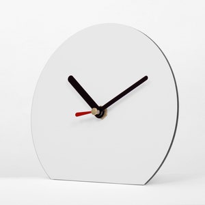 Könnte beinhalten: Eine moderne, minimalistische Uhr mit weißem Zifferblatt und schwarzen Zeigern. Der Sekundenzeiger ist rot, und die Uhr hat einen schwarzen Rand. Die Uhr hat eine halbkreisförmige Form und steht auf einer weißen Oberfläche.