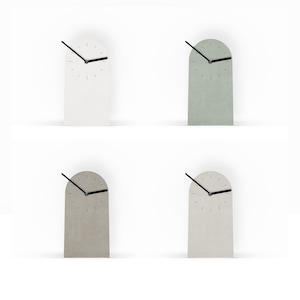 Könnte beinhalten: Vier minimalistische Schreibtischuhren in Bogenform in Weiß, Grün, Grau und Off-White. Jede Uhr hat einen schwarzen Zeiger und ein schlichtes Zifferblatt. Die Uhren werden vor einem weißen Hintergrund präsentiert.