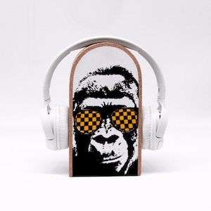 Könnte beinhalten: Ein weißer Over-Ear-Kopfhörer liegt auf einem Holzblock mit einem schwarz-weißen Gorillagesicht mit gelb-schwarz karierten Sonnenbrillen. Der Hintergrund ist schlicht weiß.