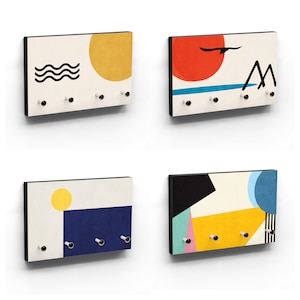 Könnte beinhalten: Vier rechteckige Schlüsselbretter mit abstrakten Kunstdesigns. Jedes hat einen schwarzen Rand, einen cremefarbenen Hintergrund und mehrere schwarze Knöpfe zum Aufhängen von Schlüsseln. Die Designs umfassen eine gelbe Sonne, schwarze Wellen, eine rote Sonne, Berge und geometrische Formen.