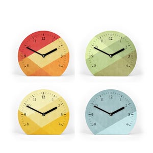 Könnte beinhalten: Vier runde Uhren mit geometrischen Mustern in verschiedenen Farben. Die Zifferblätter zeigen Farbverläufe in Rot und Orange, Grün und Olive, Gelb und Gold sowie Blau und Hellblau. Jede Uhr hat schwarze Zeiger und ein minimalistisches Design.