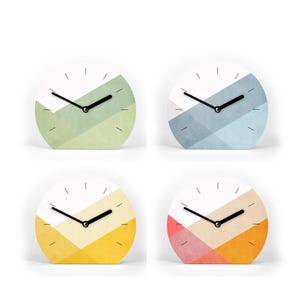 Könnte beinhalten: Vier kleine, runde Uhren mit weißen Zifferblättern und schwarzen Zeigern. Jede Uhr hat ein anderes Farbschema auf der unteren Hälfte: grün, blau, gelb und orange. Die Uhren sind vor einem weißen Hintergrund.