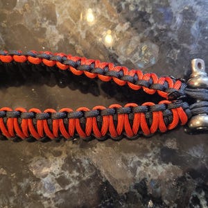 May include: A handmade paracord keychain with a stainless steel shackle. The keychain features a woven design with alternating red and black paracord. The shackle is a silver color and is attached to the paracord loop.