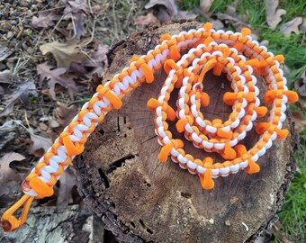 May include: A white and orange paracord bracelet with orange knots. The bracelet is coiled and lying on a tree stump.
