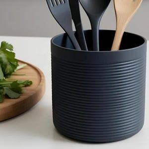 May include: A dark grey utensil holder with horizontal ridged detailing. It contains a black fork, spoon, and a wooden spoon. A wooden chopping board with green herbs is in the background.
