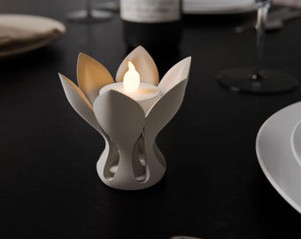 Puede incluir: Un portavelas blanco con forma de loto con una vela LED encendida. El soporte tiene lados en forma de pétalo y se asienta sobre una superficie oscura. El portavelas es un artículo decorativo.