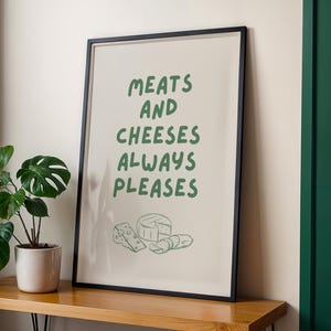 May include: A framed print with the text "MEATS AND CHEESES ALWAYS PLEASES" in green. Below the text are line drawings of cheese and meat. The print is in a black frame and sits on a wooden surface.
