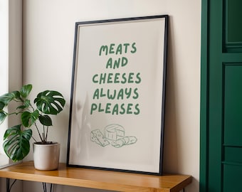 May include: A framed print with the text "MEATS AND CHEESES ALWAYS PLEASES" in green. Below the text are line drawings of cheese and meat. The print is in a black frame and sits on a wooden surface.