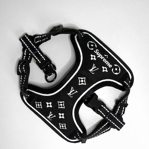 May include: Black dog harness with white trim and a white Louis Vuitton pattern. The harness has the word "Supreme" printed on it.