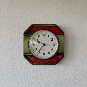 Puede incluir: Un reloj de pared vintage con esfera blanca y números negros. El reloj está rodeado por un marco octogonal verde y rojo. Las manecillas del reloj señalan las 10:30. La esfera del reloj tiene la palabra "Junghans" impresa.