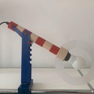 Könnte beinhalten: Eine rot-weiß-blau gestreifte Lampe mit einem blauen Sockel und einem klaren, runden Schirm. Die Lampe ist verstellbar und hat ein einzigartiges Design.