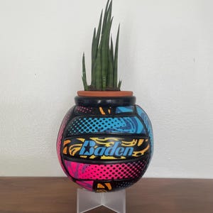 May include: A repurposed Baden volleyball planter featuring a vibrant, multi-colored pop art pattern in shades of blue, pink, yellow, and black. The ball is cut open at the top to hold a terracotta pot containing a green snake plant.