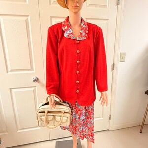 May include: A red blazer with gold buttons and a floral-print collar, paired with a matching floral skirt. A straw hat and a cream-colored handbag complete the ensemble. The outfit is displayed on a mannequin.