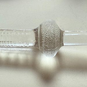 May include: A clear glass bottle stopper with a textured center and a cylindrical design. The stopper has text etched into the glass. The stopper is designed to fit into a bottle.