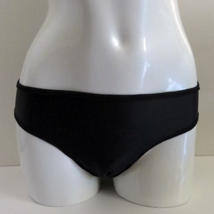 Peut inclure: Culotte noire à taille basse présentée sur un mannequin blanc. Les bords sont bordés d'une fine bordure noire. Le tissu semble lisse et opaque. Design simple et classique.