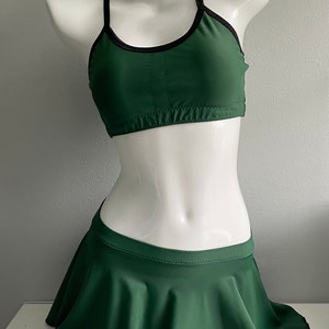 Puede incluir: Un conjunto de danza de dos piezas de color verde oscuro con ribete negro. La parte superior es de estilo halter con cuello redondo y la falda es una falda circular con un dobladillo festoneado.