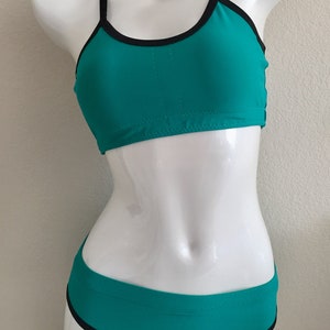 Op de afbeelding: Turquoise bikini top en broekje met zwarte bies. De top heeft een racerback-stijl en het broekje is hoog getailleerd.