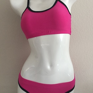 Könnte beinhalten: Ein pinkfarbener Bikini-Oberteil und -Unterteil mit schwarzem Besatz. Das Oberteil hat einen Racerback-Stil und das Unterteil ist ein frecher Schnitt.