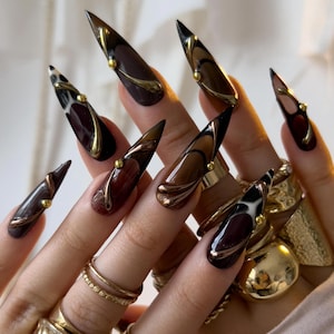 Peut inclure: Ongles artificiels en forme de stiletto, avec un design marron foncé et doré. Les ongles présentent un effet dégradé, passant du marron foncé à une teinte plus claire, avec des accents dorés et de petites perles dorées. Diverses bagues dorées ornent les doigts.