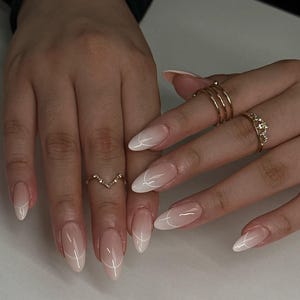 May include: Close-up of manicured nails with a French tip design. The nails are oval-shaped with a nude base and white tips, accented by a thin white line. Gold rings adorn the fingers.