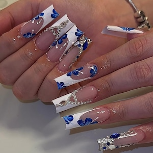 May include: Long, square-shaped artificial nails with a white base and blue floral designs. Some nails feature clear tips and are adorned with rhinestones and blue gems. The nails are long and elegant, showcasing a detailed manicure.