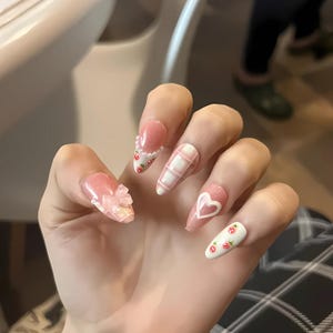 May include: A hand displaying long, almond-shaped artificial nails. The nails are decorated with pink glitter, a white and pink checked pattern, a white heart outline, and small strawberry designs. One nail has a pink bow.