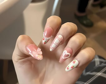 May include: A hand displaying long, almond-shaped artificial nails. The nails are decorated with pink glitter, a white and pink checked pattern, a white heart outline, and small strawberry designs. One nail has a pink bow.