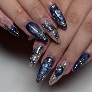 May include: A set of long, almond-shaped artificial nails. The nails feature a dark blue, glittery base with silver embellishments, including crescent moons, butterfly-like shapes, and celestial designs. Some nails have a clear base with silver accents.