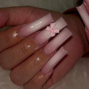 Op de afbeelding: Lange, vierkante acrylnagels met een gradiënteffect, overgaand van zachtroze naar wit. Eén nagel heeft een kleine, lichtroze strik. De nagels zijn glanzend en goed verzorgd, wat een modern nail art design laat zien.