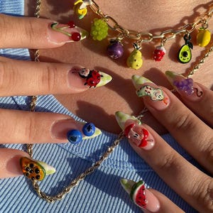 May include: Close-up of a gold necklace with fruit charms and long, decorated fingernails. The nails feature 3D fruit designs, including cherries, ladybugs, grapes, blueberries, and watermelon slices. The necklace has charms of a strawberry, avocado, and other fruits.