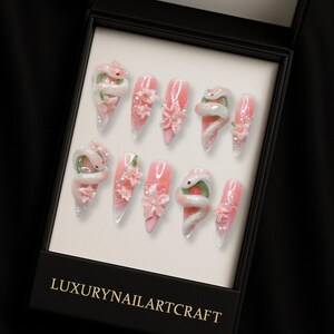 May include: A set of ten artificial nails in a black box. The nails are pink with white tips and decorated with white snakes, pink flowers, and glitter. The box has the text "LUXURYNAILARTCRAFT" in gold.