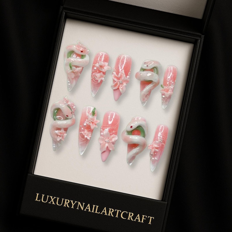 May include: A set of ten artificial nails in a black box. The nails are pink with white tips and decorated with white snakes, pink flowers, and glitter. The box has the text "LUXURYNAILARTCRAFT" in gold.