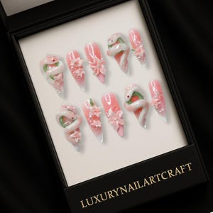 May include: A set of ten long, almond-shaped artificial nails. Each nail features a gradient pink and white design, adorned with 3D snake and floral embellishments. The nails are presented in a black box with the text "LUXURYNAILARTCRAFT".