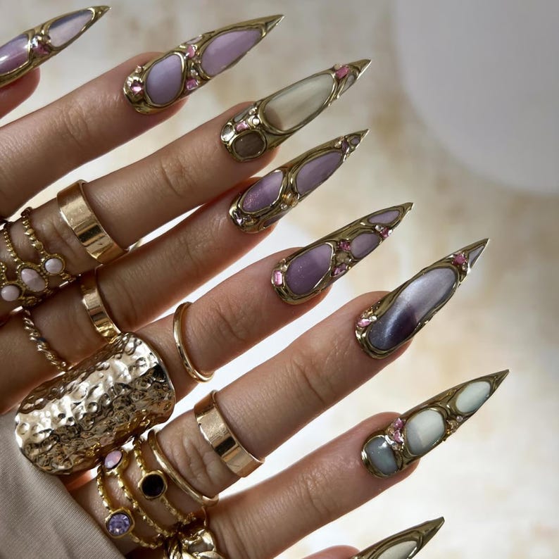 May include: Close-up of long, pointed fingernails adorned with gold-colored metal and various pastel-colored stones. The fingers are also decorated with multiple gold rings of different styles and sizes, creating a glamorous aesthetic.