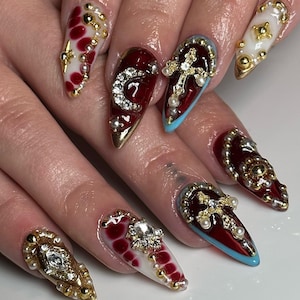 Op de afbeelding: Een set lange, puntige nagels met uitgebreide versieringen. De nagels tonen diverse ontwerpen, waaronder rode en witte patronen, gouden details, parels en glinsterende edelstenen. Sommige nagels hebben een kruismotief, terwijl andere een met juwelen bezette letter "C" hebben.